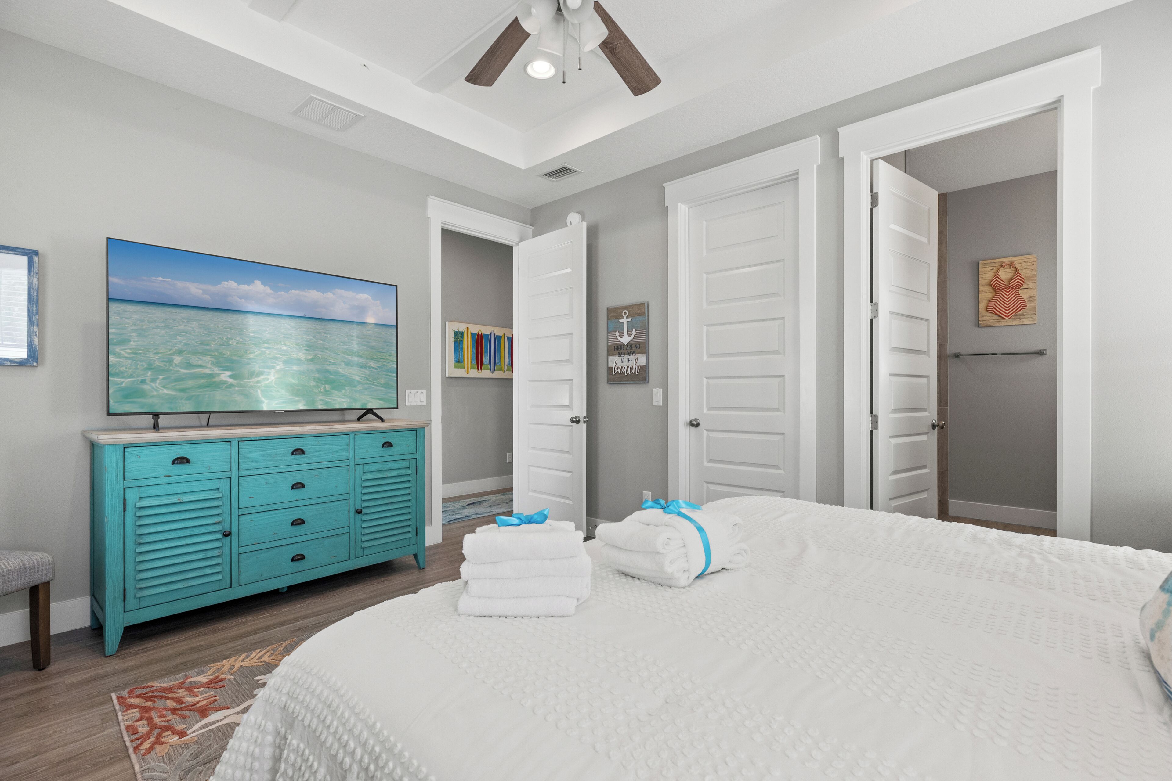 Une chambre avec un grand lit, une commode, un ventilateur de plafond et un tableau représentant l’océan.