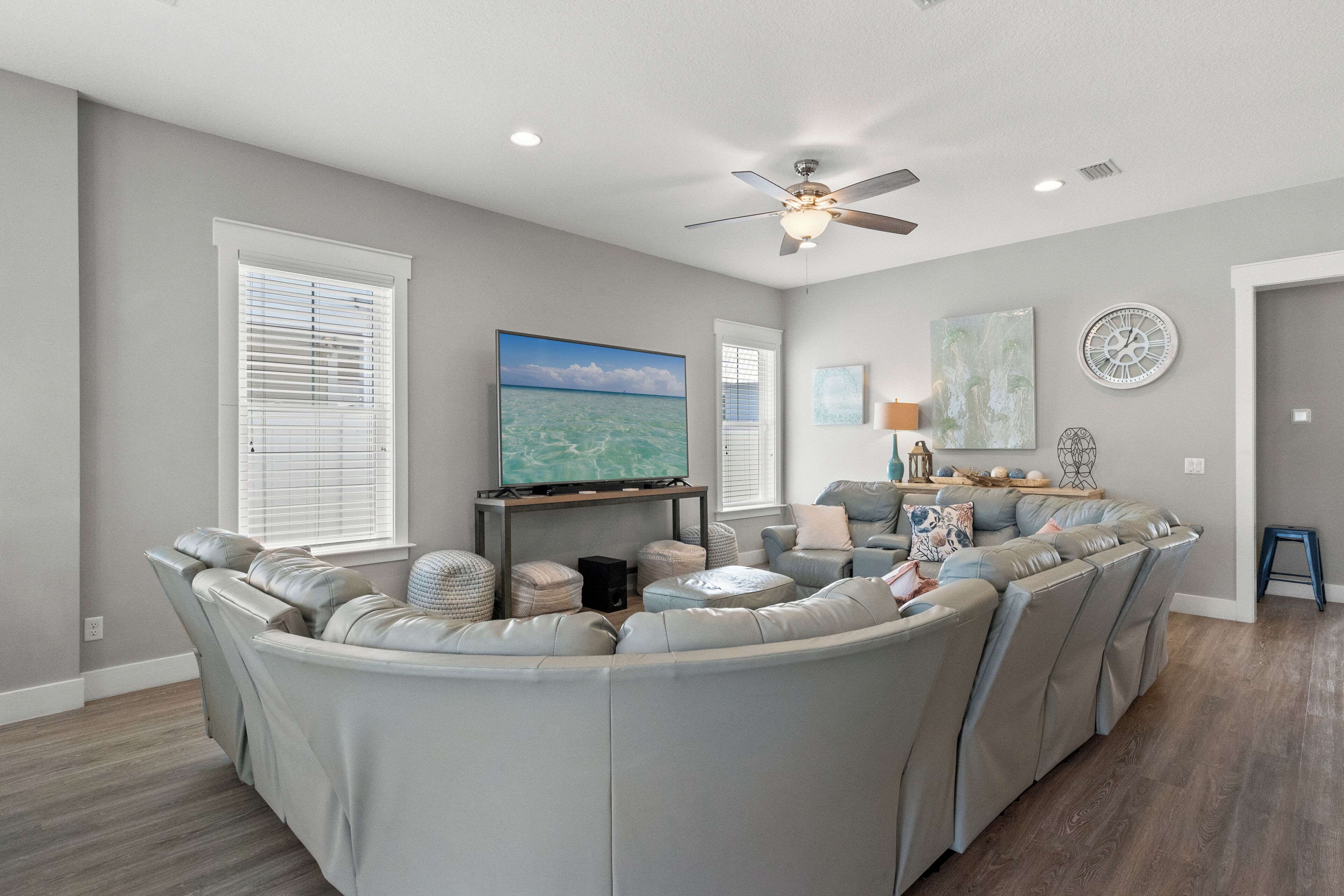 A living room with a large L-shaped sectional sofa, a ceiling fan, a flat-screen TV, and a large window with blinds.