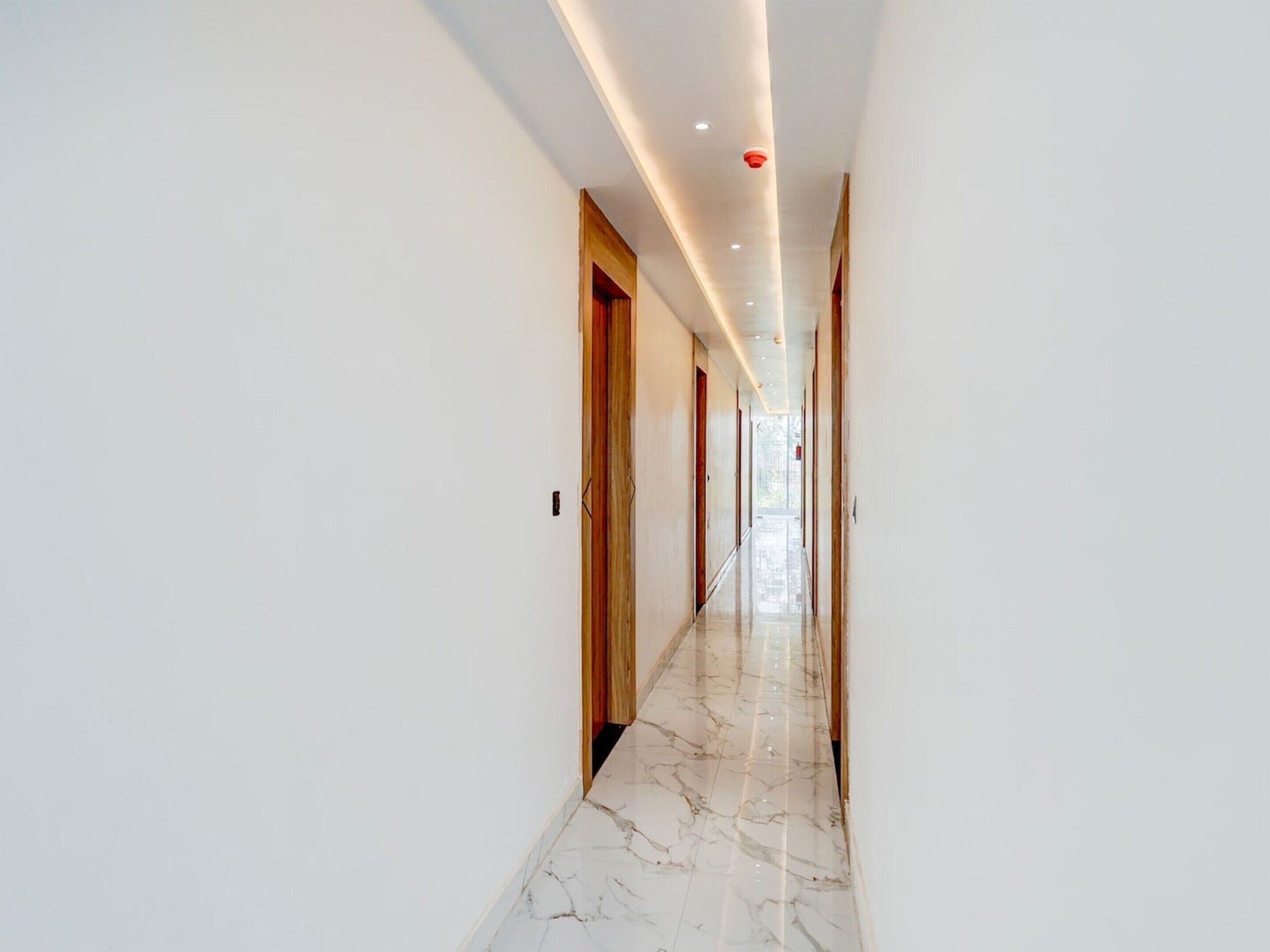 A long, narrow hallway with white walls and a marble floor, leading to a bright room at the end.