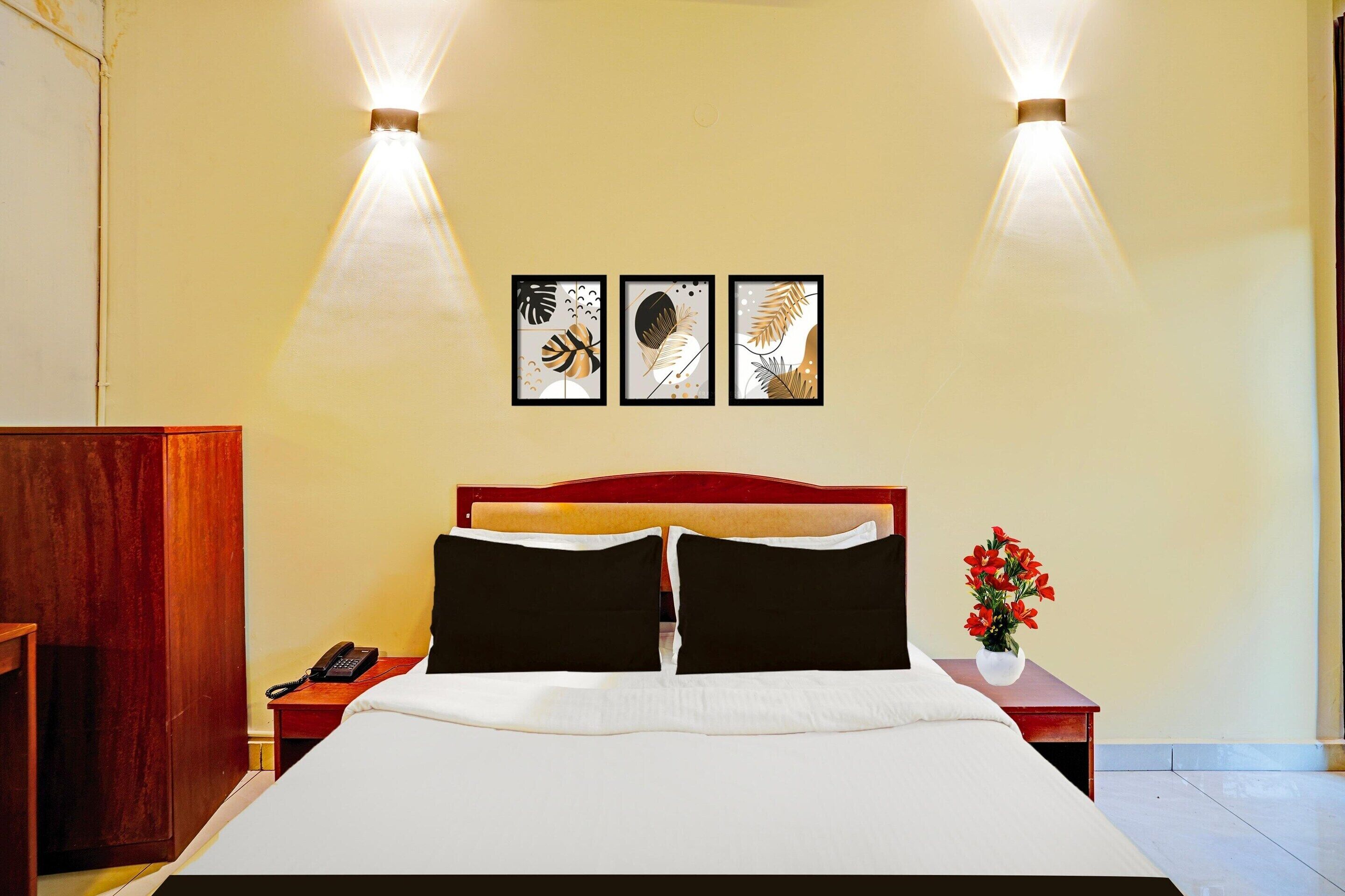 A hotel room with a bed, a nightstand with a phone, a vase with flowers, and three framed pictures on the wall.