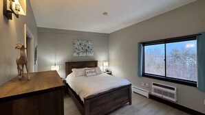 A bedroom with a wooden bed, a nightstand, a wall-mounted lamp, a decorative deer, a floral artwork, and a window with a view of trees. - Walk to Weirs Beach Boardwalk with Treetop Views of Lake Winnipesaukee! (Laconia)
