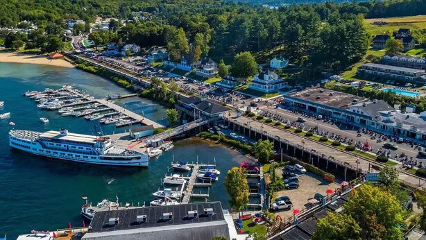 A marina with boats docked, a pier, and buildings with visible windows and storefronts. - Walk to Weirs Beach Boardwalk with Treetop Views of Lake Winnipesaukee! (Laconia)