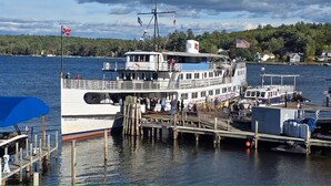 A multi-deck boat docked at a pier with a view of a forested shoreline. - Walk to Weirs Beach Boardwalk with Treetop Views of Lake Winnipesaukee! (Laconia)