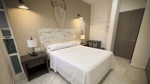 A hotel room with a bed, bedside tables, lamps, a decorative wall hanging, and a door. - Beachside Nook - Modern Oceanfront Condo, Steps from the Beach with Pool (Cozumel)