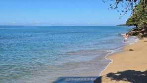 Une plage à l’eau claire, une personne qui se promène et des affleurements rocheux.