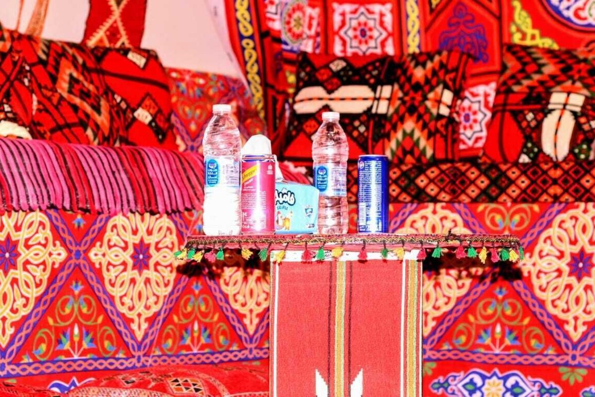 Une pièce dotée d’un tapis rouge à motifs, d’une petite table sur laquelle sont posées des bouteilles d’eau, et d’une bordure décorative.