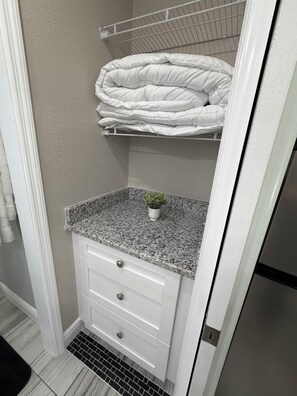 Un baño con encimera de granito, una pequeña planta en maceta, un mueble blanco con cajones y un perchero blanco en la parte superior.