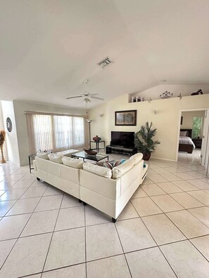 A spacious living room with a white sofa, a flat-screen TV, a ceiling fan, and a large window with blinds. - Villa Blue Water - Cape Coral (Cape Coral)