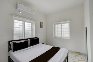 A bedroom with a bed, two windows, an air conditioning unit, and a small bedside table. - Deluxegn Luxury Suits (Hyderabad)