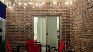 A brick-walled patio with a glass door, red chairs, a table, and string lights.