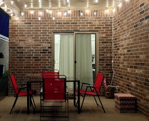 Un patio con paredes de ladrillo, puerta de vidrio, sillas rojas, una mesa y luces colgantes.