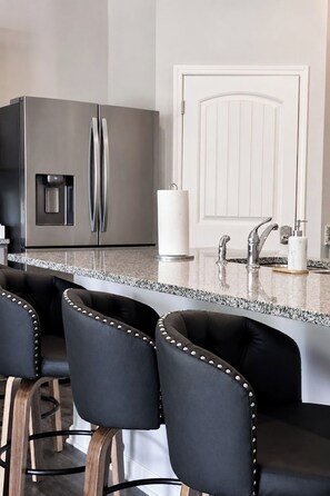 Una cocina con un refrigerador de acero inoxidable, un dispensador de agua, una puerta blanca, un lavamanos con grifo y taburetes de bar con patas de metal.