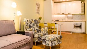 Een woonkamer met een bank, een fauteuil met een patroon, een kleine tafel met stoelen, en een keuken met kasten en apparatuur.