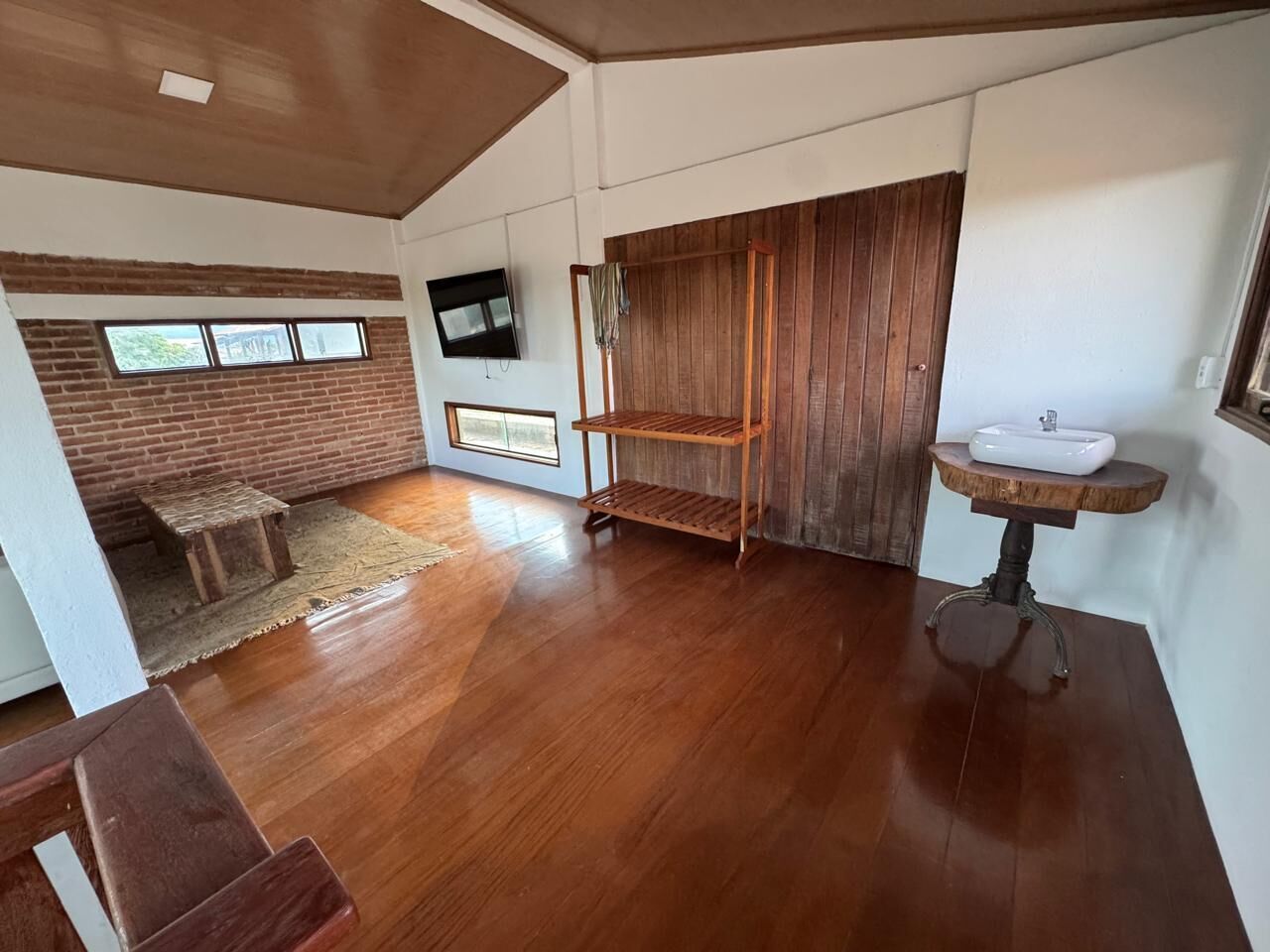 A room with a wooden floor, a TV mounted on the wall, a wooden bench, a small table with a sink, and a window with a view of the outside.