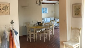 Een eetgedeelte met een tegelvloer, witte muren en een houten plafond. Er staan twee witte stoelen, een tafel met een laptop en een kroonluchter.