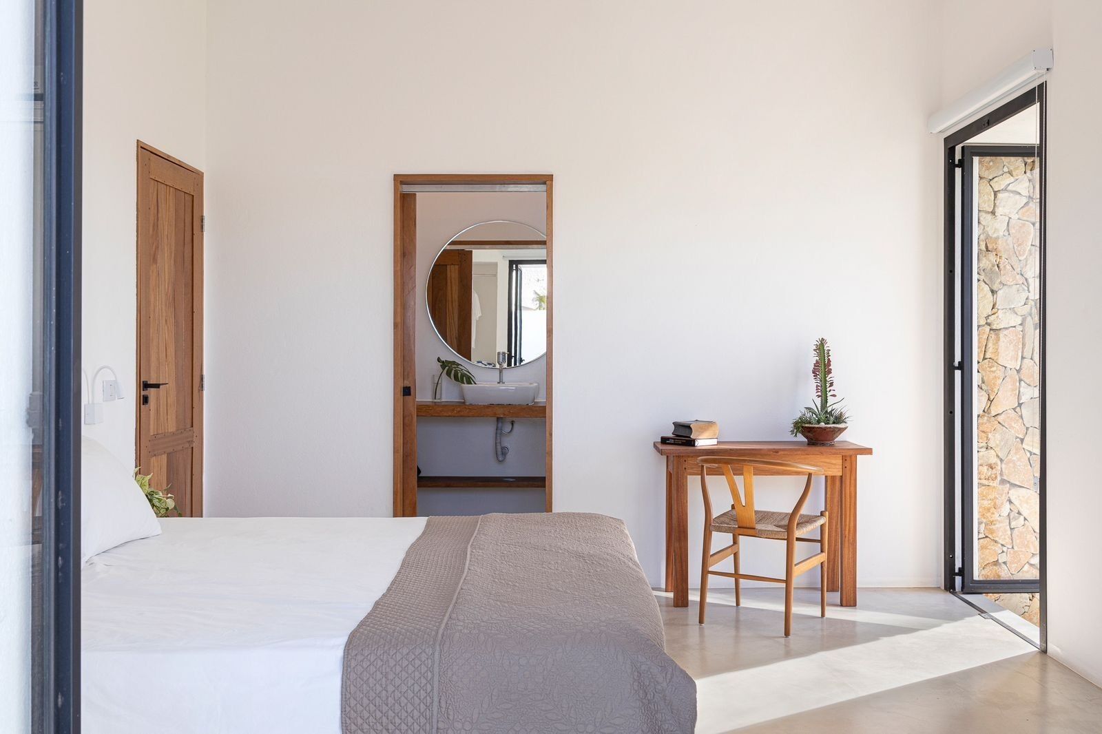 A bedroom with a bed, a wooden desk, a chair, a mirror, and a bathroom visible through an open door.