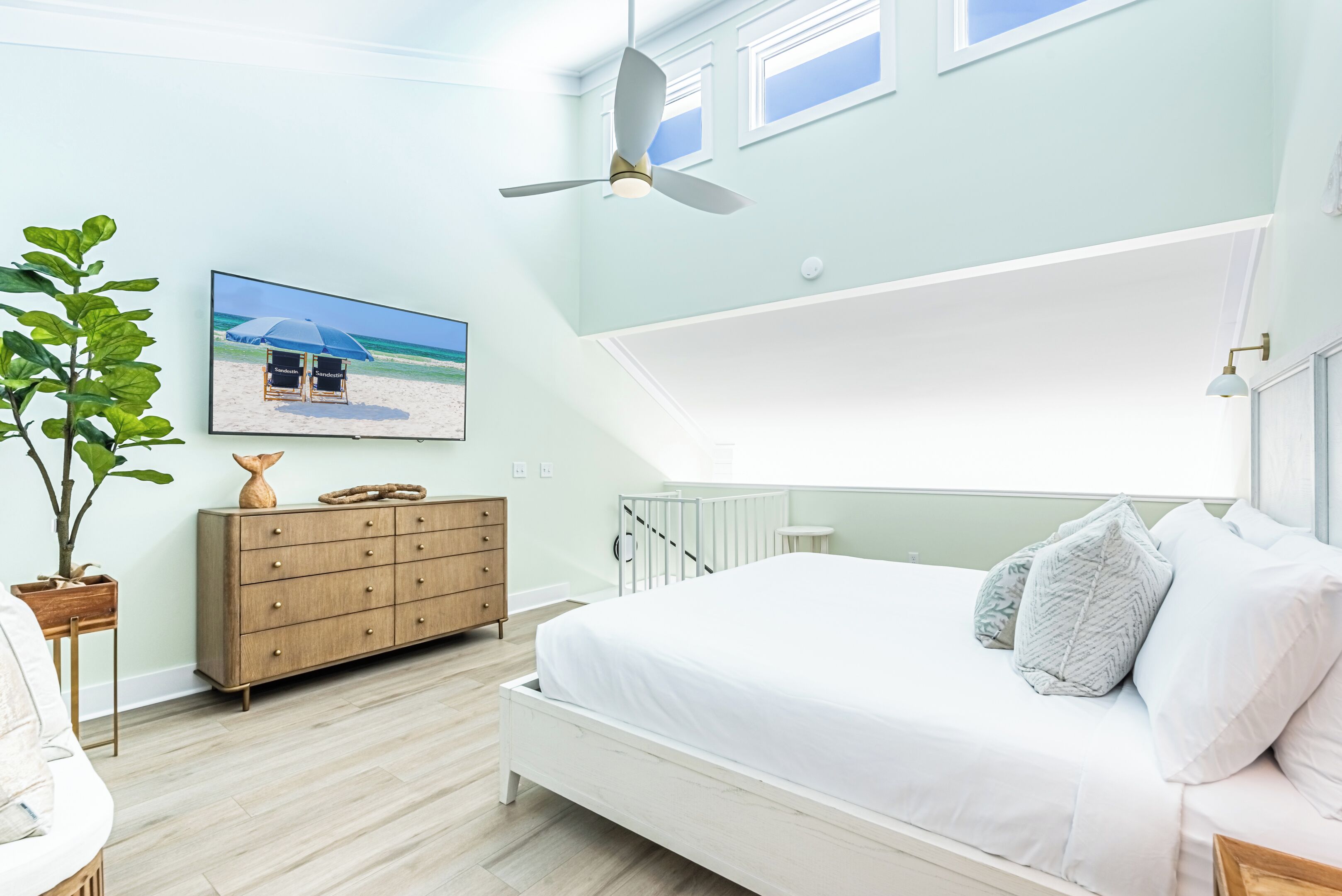 Une chambre à coucher avec un lit, un ventilateur de plafond, une commode, une plante et un tableau sur le thème de la plage accroché au mur.