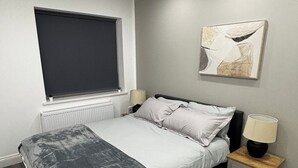 A bedroom with a bed, bedside table, lamp, window, radiator, and a wall-mounted abstract painting. - Cosy 2 Bed-5 Guests-garden-parking&wifi (London)