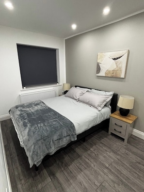 A bedroom with a bed, bedside table, lamp, window, radiator, and a wall-mounted abstract painting. - Cosy 2 Bed-5 Guests-garden-parking&wifi (London)