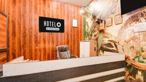 A hotel reception desk with a wooden wall, a sign reading 'HOTELO,' a fan, and a decorative wall mural. - Deluxehotel Mayur Residency (Gorakhpur)