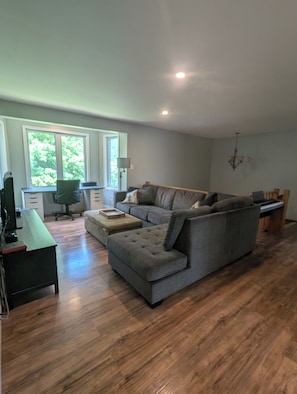Een moderne woonkamer met een grijze modulaire bank, een houten salontafel, een bureau met een computer, en een raam met uitzicht op bomen.