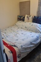 A bed with a nautical-themed bedspread, pillows with a Union Jack design, and a plain wall.