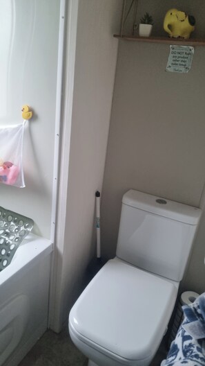 A bathroom with a toilet, a white sink, a shower curtain, a shelf with a plush toy, and a sign instructing not to flush anything other than toilet tissue. - Villa With Decking & Parking (Clacton-on-Sea)