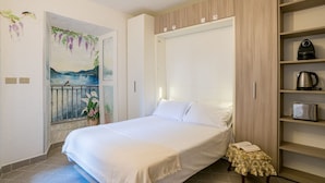 Un dormitorio con cama, una mesita de noche con un libro, un estante con electrodomésticos de cocina y un cuadro en la pared.