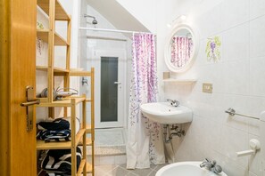 A bathroom with a wooden shelf, a shower curtain, a sink, and a mirror. - Cozy studio apartment of approximately 30 square meters, perfect for two people, located on the ground floor of a building (accessible via a few steps).The property is located in Argegno, a charming village on the western shore of Lake Como, in a well-se (Argegno)