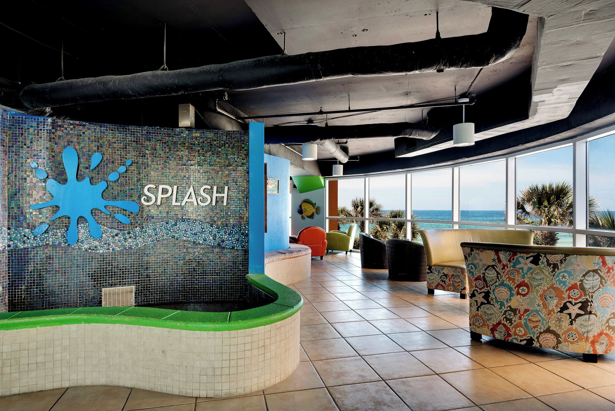 A modern indoor pool area with a large blue splash mural, a seating area with colorful chairs, and a view of the ocean through large windows.