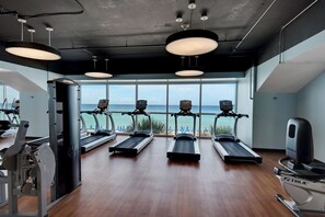 Een fitnessruimte met loopbanden, een roeimachine en een gewichtbank. Grote ramen bieden uitzicht op de oceaan.