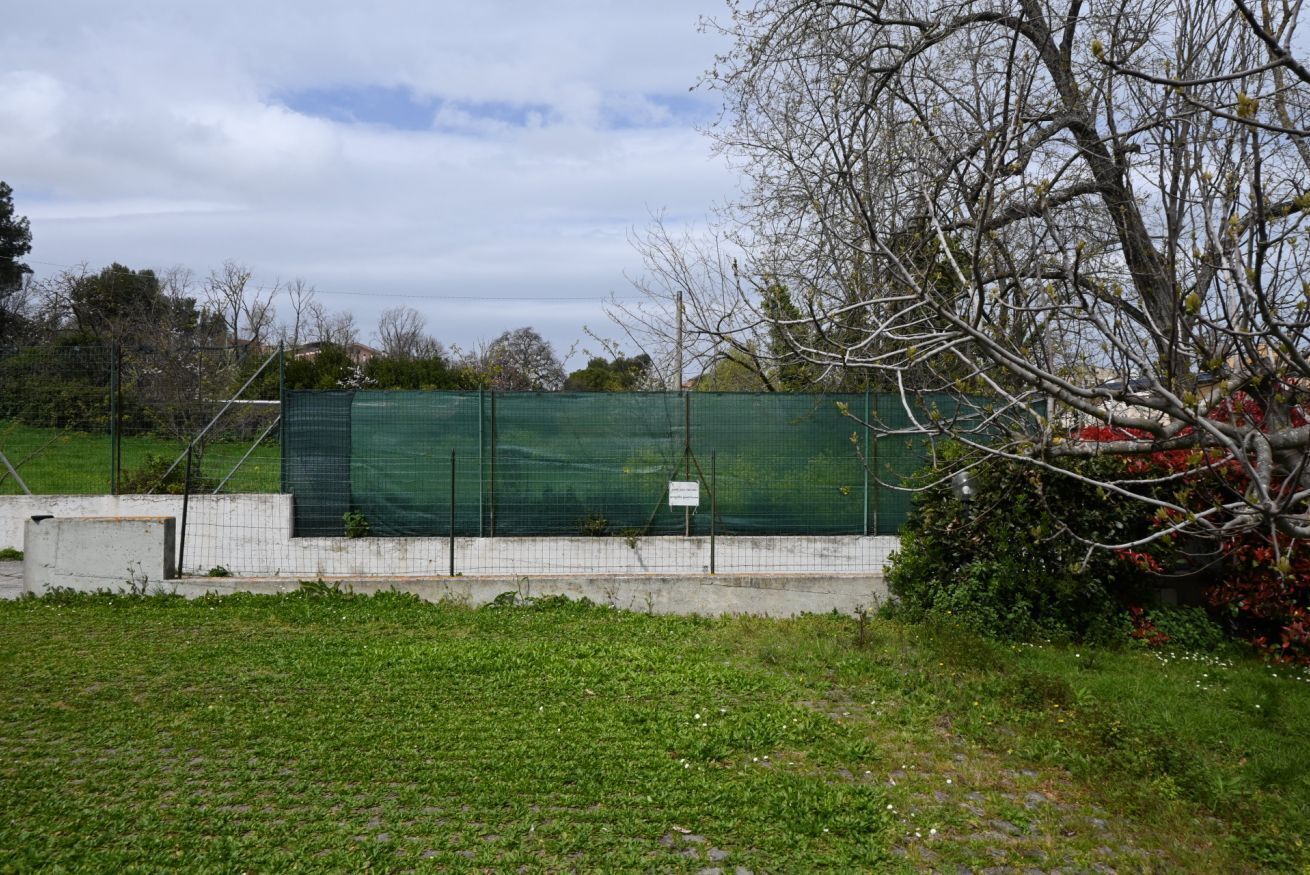 A fenced area with a green netting structure, a concrete wall, and a grassy field.