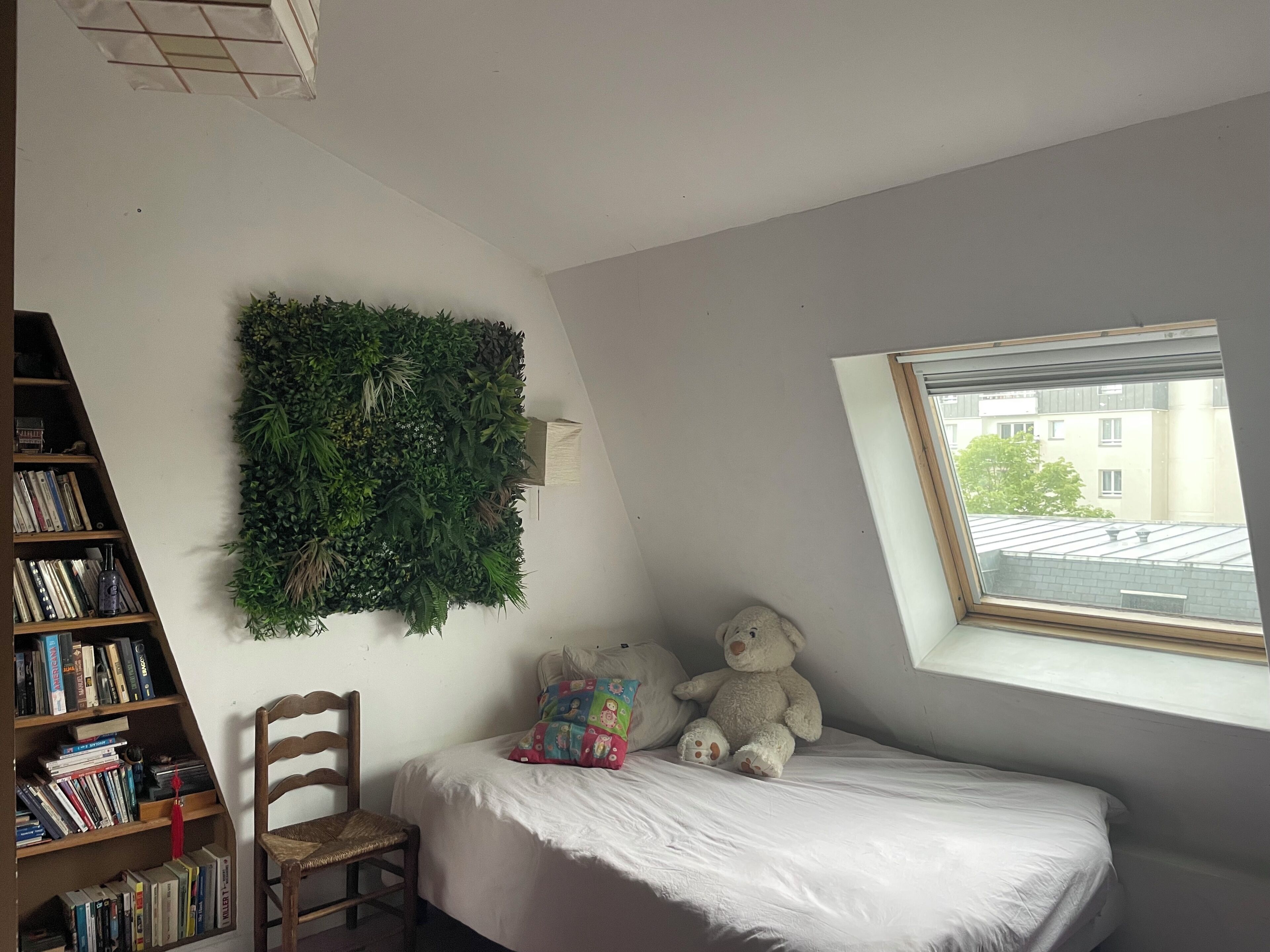Une chambre à coucher avec un lit, une chaise, une bibliothèque, une plante fixée au mur, un ours en peluche et une fenêtre.