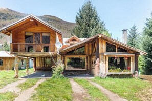 A wooden cabin with a balcony, a covered porch, and a mountain backdrop.