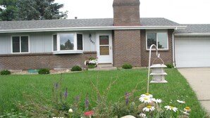 Una casa residencial con fachada de ladrillo, un jardín delantero con césped y flores, y un garaje.