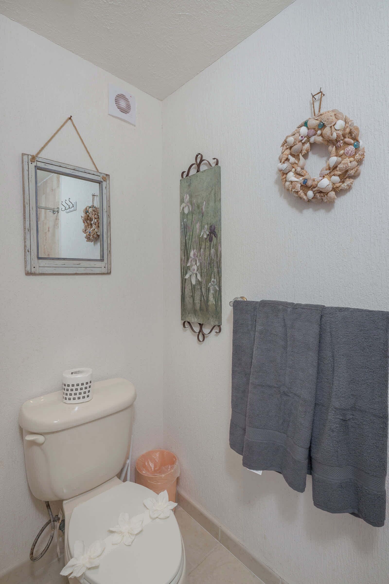 Et badeværelse med toilet, skraldespand, håndklædestang med håndklæde, indrammet spejl og en dekorativ krans på væggen.