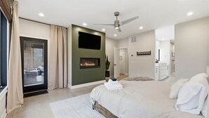 Ein modernes Schlafzimmer mit einem großen Bett, einem Deckenventilator, Einbauleuchten, einem Kamin und einer Glastür, die auf einen Balkon führt.