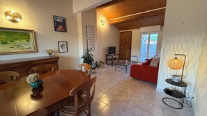 Uma área de refeições com mesa e cadeiras de madeira, uma TV em um suporte e um sofá vermelho ao fundo.