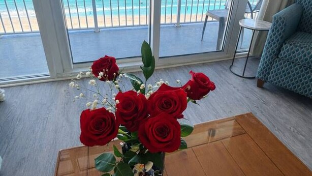 Una habitación con un ventanal que da a una playa, una mesa de madera con un jarrón de rosas rojas y un sofá azul con estampado.