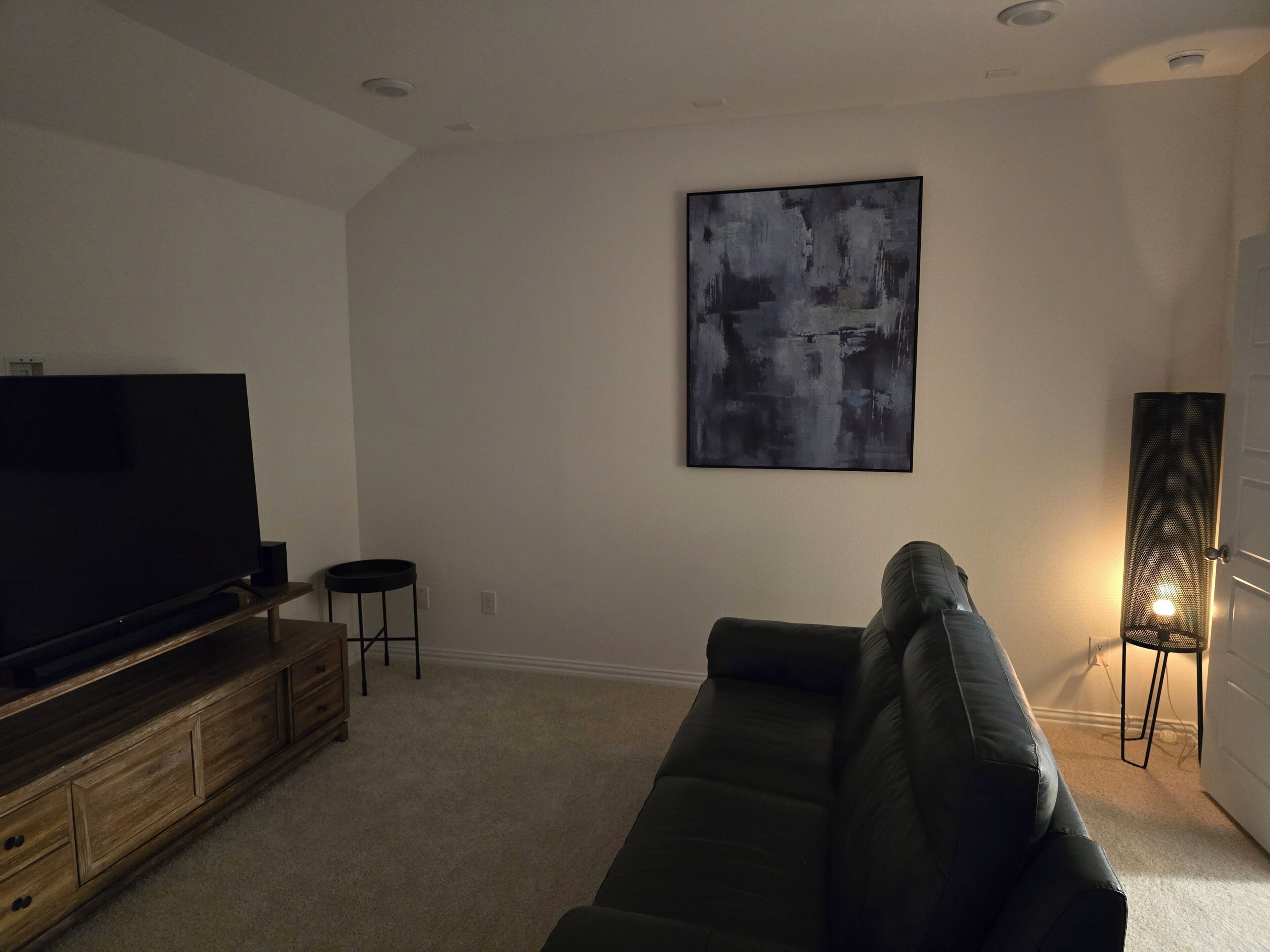 A living room with a flat-screen TV on a wooden stand, a black leather sofa, a black abstract painting on the wall, and a floor lamp.