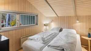 Een kamer met houten wanden, een bed met wit beddengoed, een raam met zonwering, een nachtkastje met een lamp en een radiator.