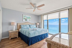 Une chambre à coucher comprenant un lit, deux tables de chevet, un ventilateur de plafond, une grande fenêtre avec vue et un fauteuil en osier.