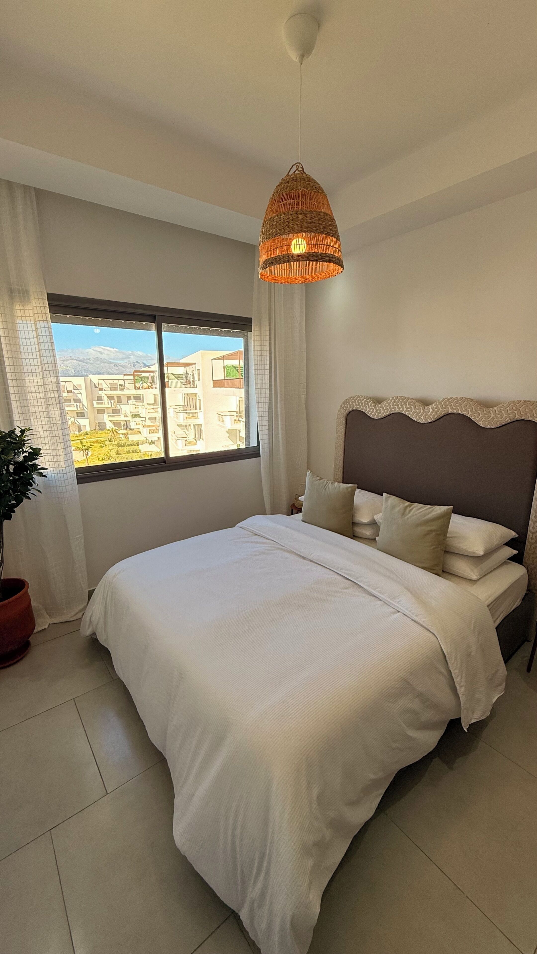Ein Schlafzimmer mit einem großen Bett, einer hängenden Korbschwinge, Blick auf Gebäude durch ein Fenster und einer Zimmerpflanze.