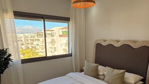 Une chambre avec un grand lit, un luminaire suspendu en osier, une vue sur les immeubles par la fenêtre et une plante en pot.