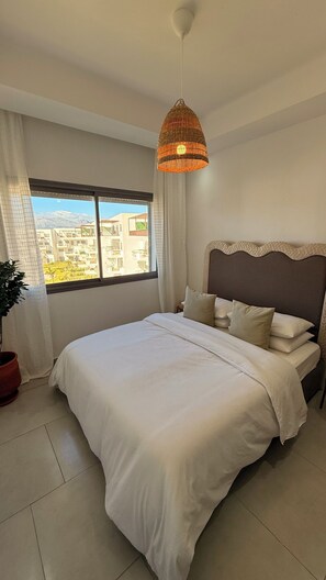Ein Schlafzimmer mit einem großen Bett, einer hängenden Korbschwinge, Blick auf Gebäude durch ein Fenster und einer Zimmerpflanze.