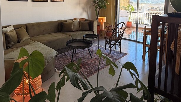 Un salon comprenant un canapé, une table, des chaises, un luminaire suspendu et une grande fenêtre donnant sur un beau paysage.