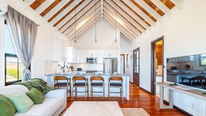 Una cocina moderna con armarios blancos, una isla central con taburetes de bar, un televisor grande y un área de comedor con un sofá blanco y cojines verdes.