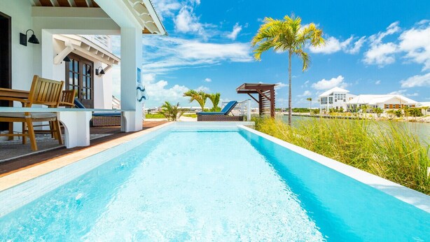 Una piscina con sillones de descanso, una terraza de madera y vistas a la costa con casas y palmeras.