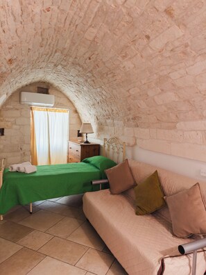 A room with a stone vaulted ceiling, a bed with green bedding, a wooden nightstand, a lamp, and a beige sofa with pillows. - Barocco Art House - Elegant apartment with terrace in Martina Franca (Martina Franca)