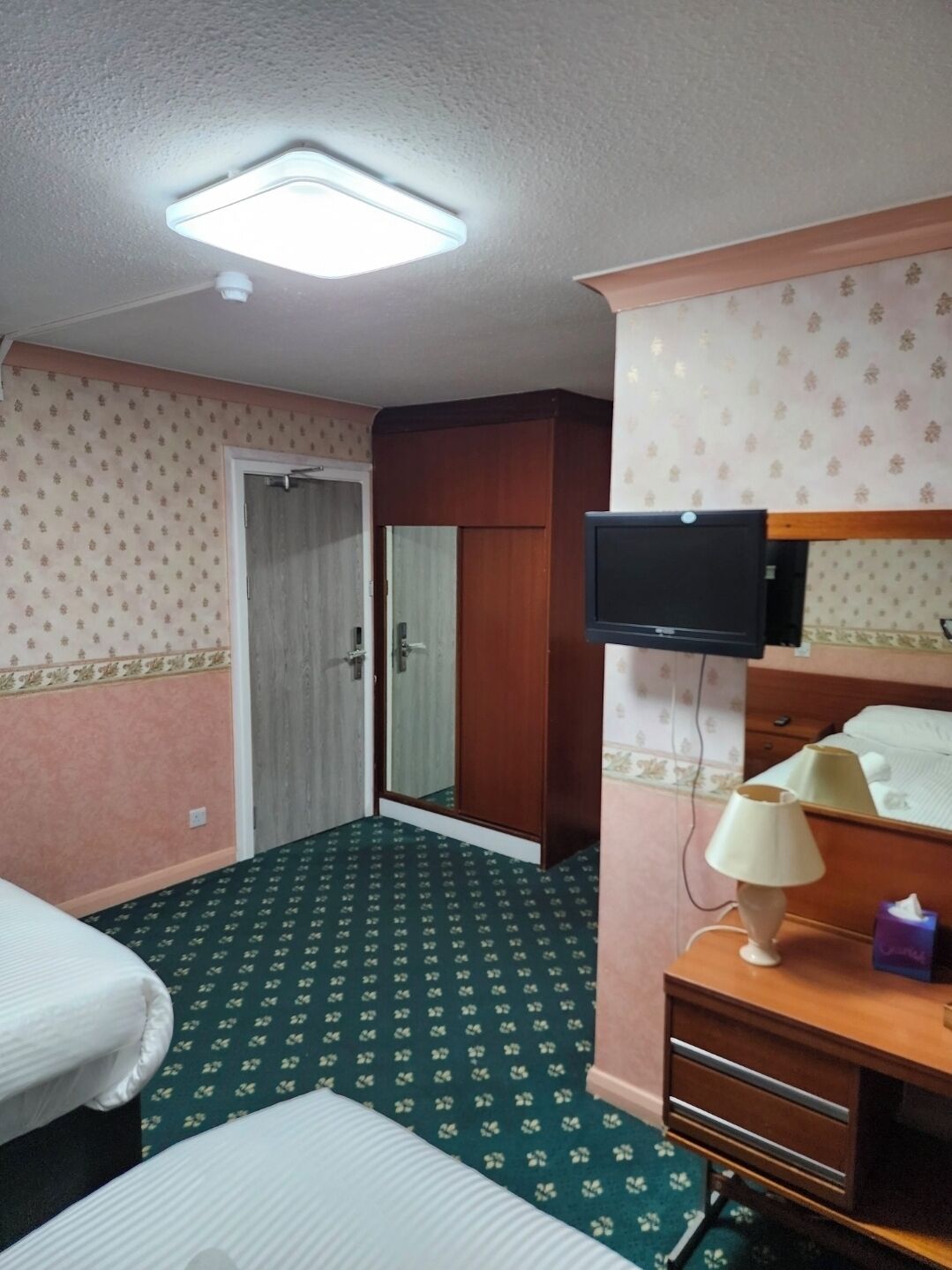 Quarto de hotel com uma cama, escrivaninha com abajur, televisão e guarda-roupa.